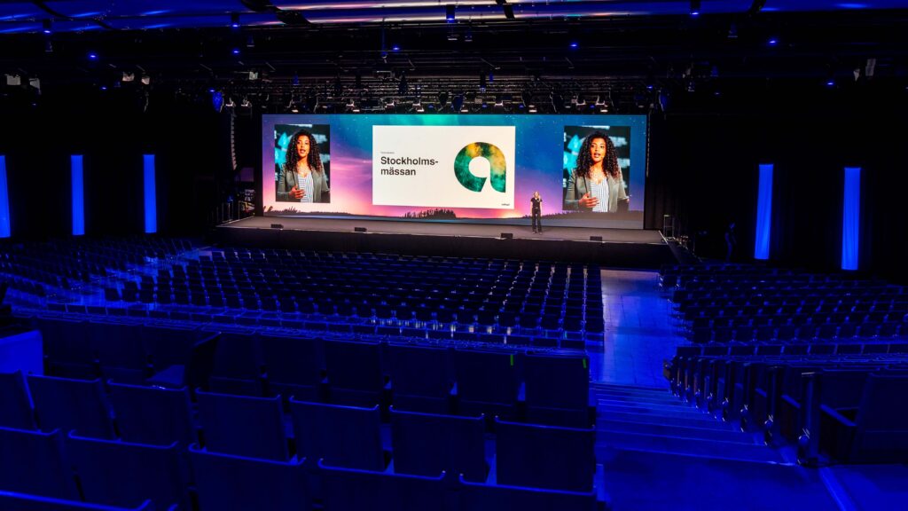 A large, modern auditorium with rows of empty seats facing a brightly lit stage. A presenter stands on stage near a screen displaying Stockholmsmässan and colorful graphics, with two large portraits on either side.