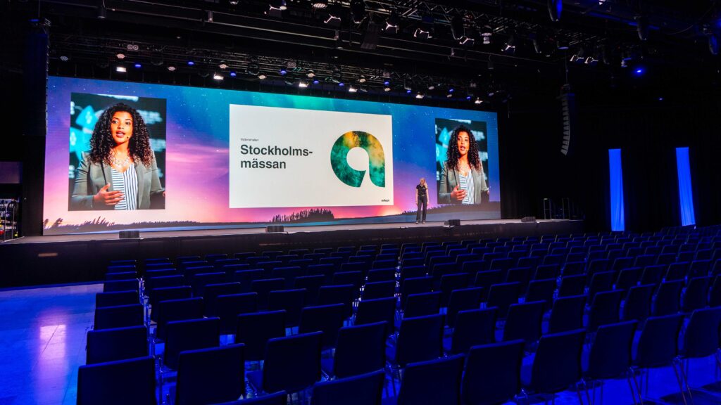 A large conference hall with rows of empty chairs faces a stage where a person stands. Two large screens display a woman speaking and the text “Stockholmsmässan” with a circular green-blue logo.