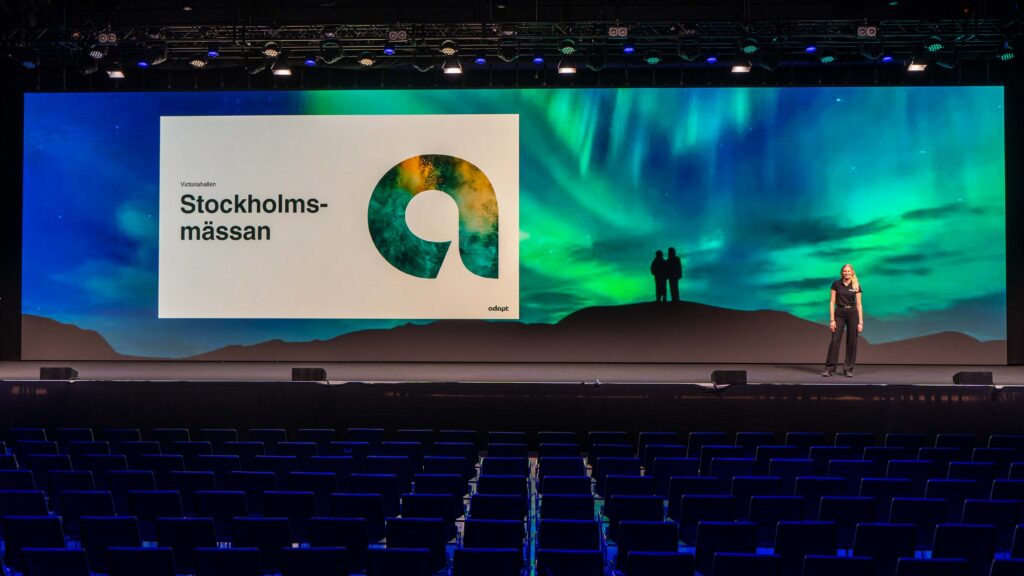 A woman stands on a stage with a large screen behind her displaying Stockholmsmässan and colorful graphics. The backdrop shows a digital image of a northern lights landscape with two silhouetted figures. Rows of empty seats are visible.
